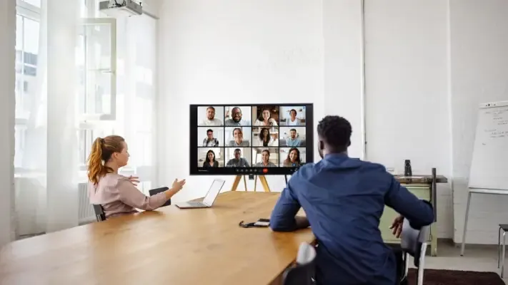 Dois profissionais em uma sala de reunião moderna participando de uma videoconferência com uma equipe remota em uma tela grande.